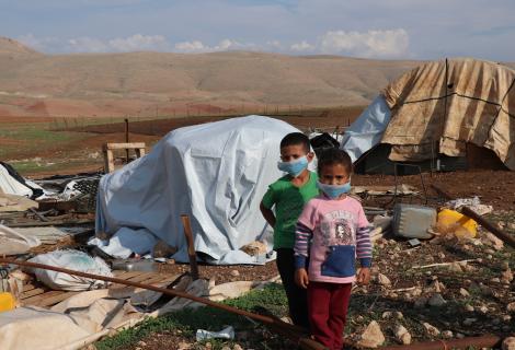 A little boy and a little girl wearing facemasks stand in front of tents destroyed by Israeli forces in Palestine