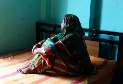 A woman sitting on a bed faces away from the camera, with her hands around her knees. 