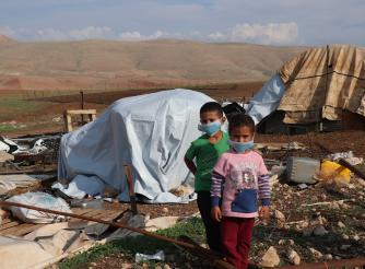 A little boy and a little girl wearing facemasks stand in front of tents destroyed by Israeli forces in Palestine